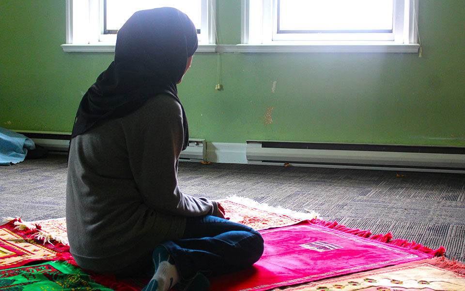 A woman in a hijab sits on a prayer mat in a women's prayer space.